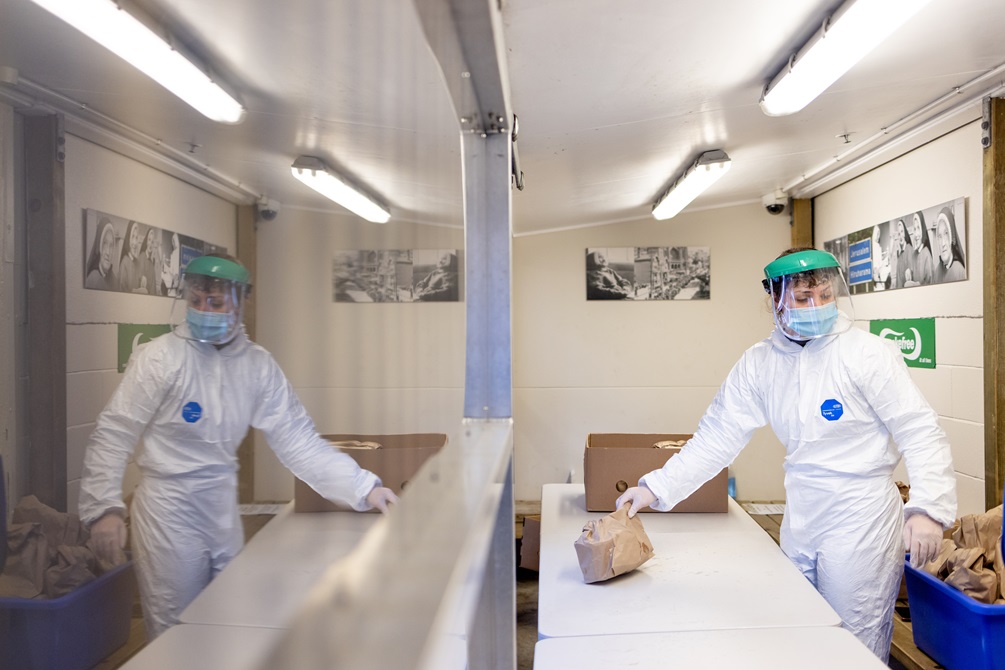 A person wearing protective clothing and a protective mask on, holding onto a brown paper bag in a food preparation site.