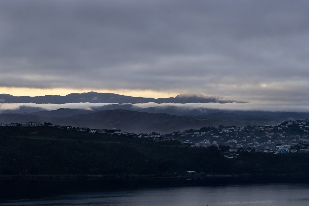 Wellington hills with houses dotted over one side. Still water and some light peeking through the clouds.