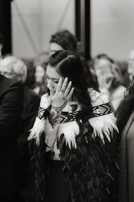 A young woman dressed in a special korowai cloak. Her hand, which is tattooed in Māori design, is brushing her dark hair from her face.