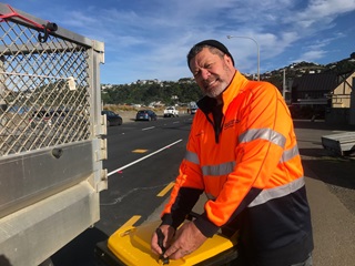Man wearing a high vis orange jacket putting a bin clip on a wheelie bin on the side of the road.