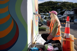 Woman smiling wearing a high vis orange jacket, painting a mural on a small building.