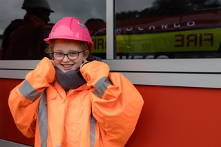 A young girl smiling at the camera wearing an orange high vis jacket and a pink hard hat.
