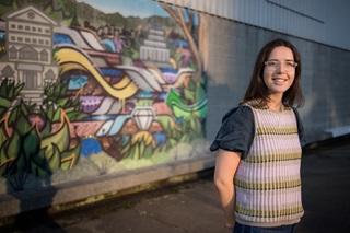 A woman wearing a colourful vest and a puffy sleeved navy top smiling infront of a mural.