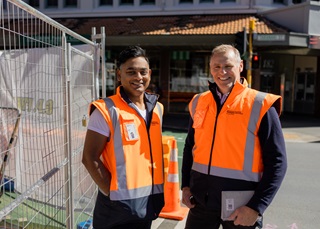 Two men wearing orange high-vis vests smiling next to each other with their hands in their pockets, standing on the road next to metal fencing.