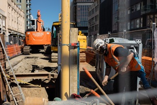 A person wearing a hard hat and high-vis vest digging into the ground with a tool on a roading worksite, which has heavy machinery and equipment.