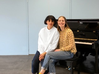 Two women sitting together at a piano. One is wearing a white shirt and the other is wearing a bright orange blouse.