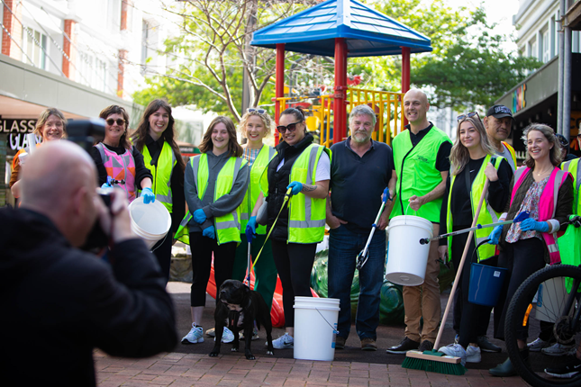About a dozen people standing in a line wearing yellow high-vis vests and gloves, holding cleaning buckets and sticks for picking up rubbish.