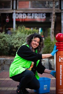 A young woman with dark curly hair and wearing a high-vis yellow vest, smiling as she crouches beside a red post, which she scrubs with a bucket of soapy water.