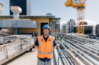 Man in a an orange high vis with a blue hard hat standing infront of a crane.