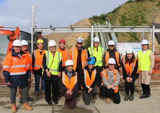 A group of women wearing orange high-vis vests and hard hats standing in two rows smiling. They are on a construction site with large pipes and a hill behind them.