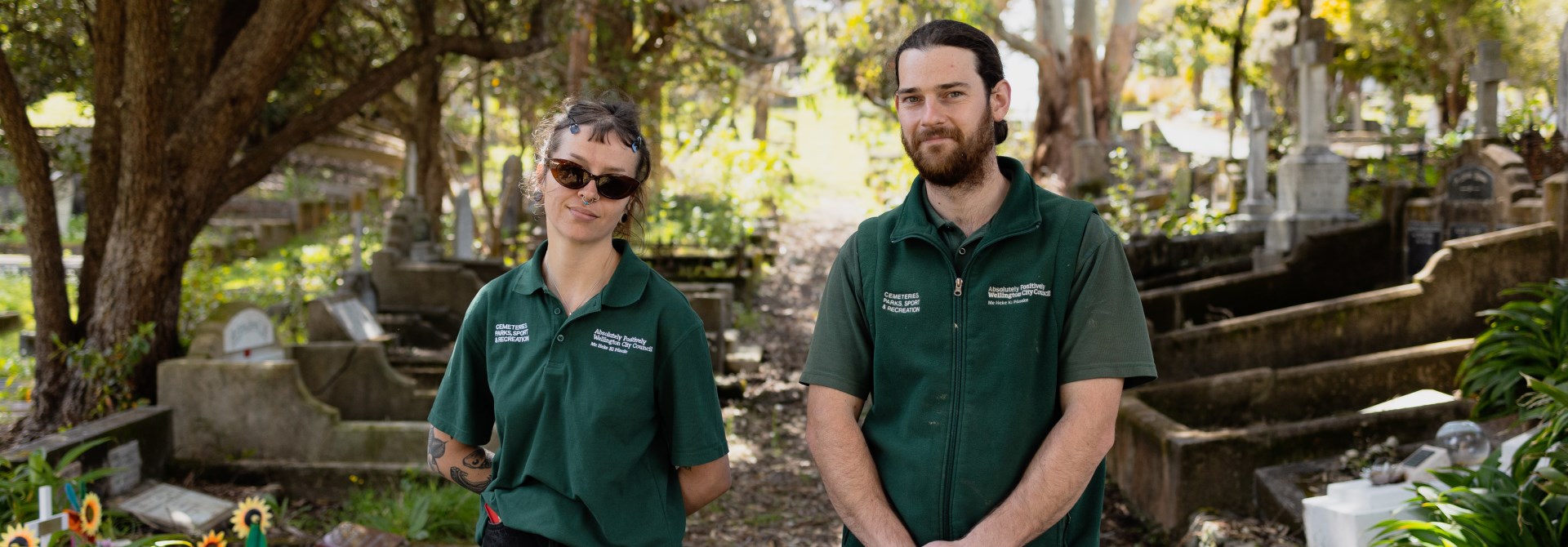 Taylor and Jayden stand in their green polo shirt uniforms under the trees in a cemetery.