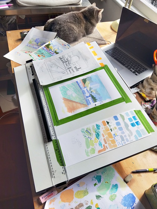 A desk with a dark grey cat, open laptop, and watercolour paintings on it.