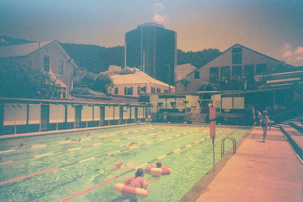 A photo of Thorndon Pool taken on Redscale film by Wellington artist Hannah Webster. There are buildings and hills in the background and people swimming in the pool.