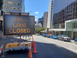 A digital road sign saying 'Shell Lane closed' surrounded by road cones on a city street with parked cars and high-rise buildings.