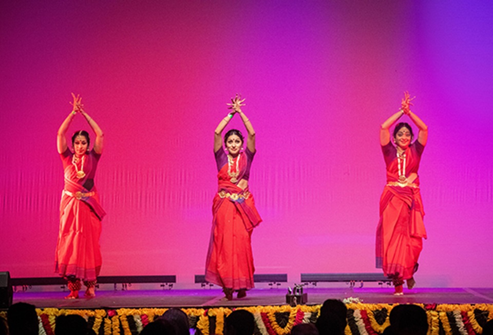 Three dancers in red dresses on stage with their hands raised above their heads.