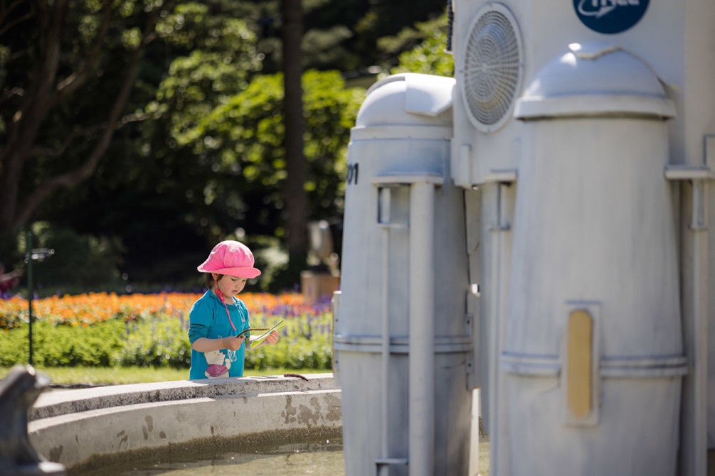 A small child looks at a sculpture of a space rocket in a garden.