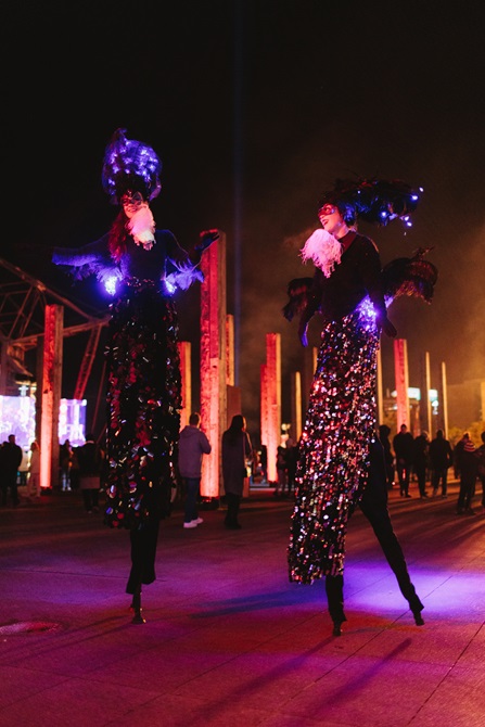 Two performers on stilts, adorned with lights and feathers.
