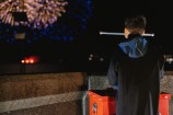 A young boy stands next to an explosives plunger while fireworks go off in the background.
