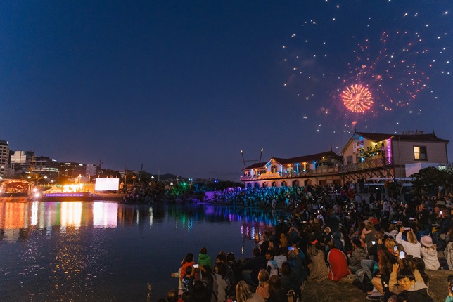 A crowd of people enjoy a concert at a waterfront lagoon, while fireworks explode above a boat shed in the background.