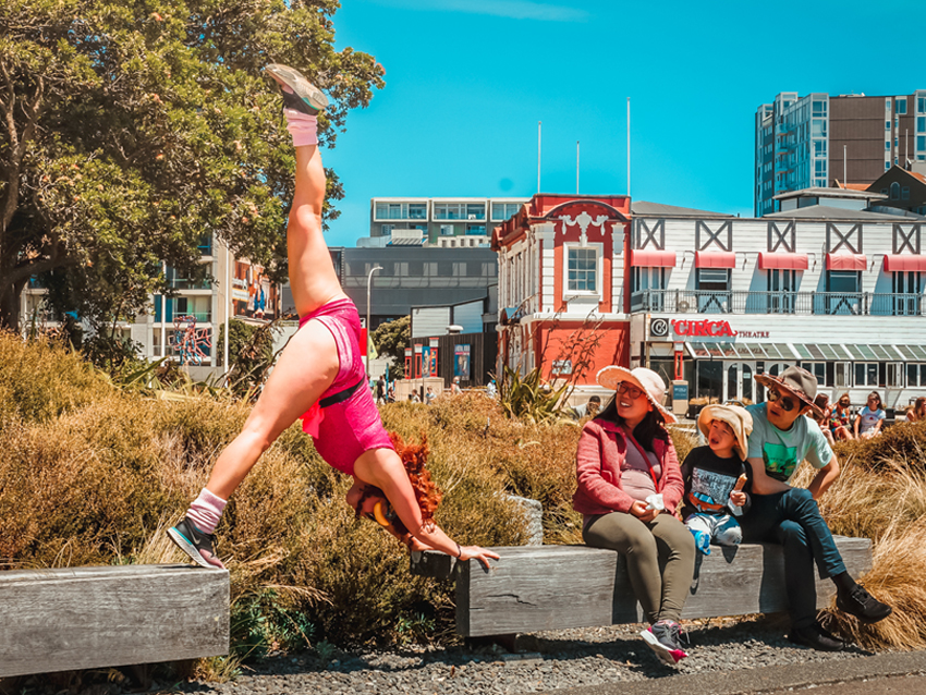 A performer doing the splits on the waterfront while a family looks on.