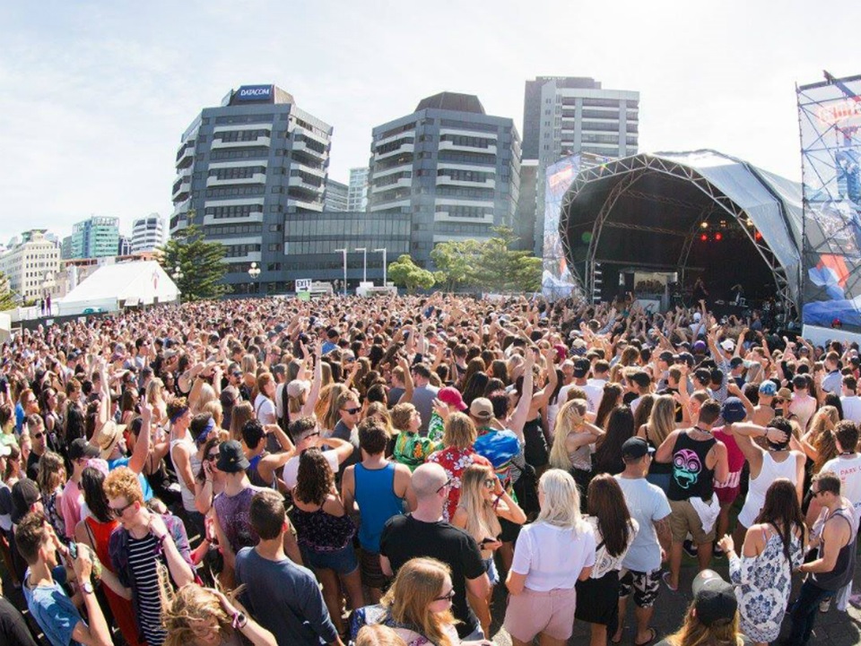 A large crowd of people dancing in front of a stage, with the city in the background.