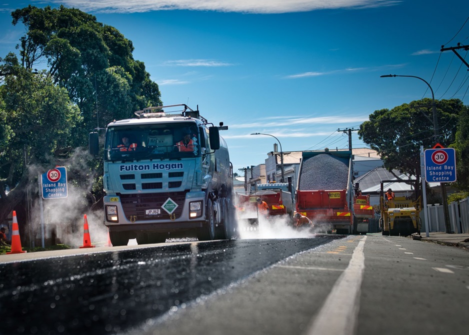 A layer of fresh bitumen on a road surface. A work crew and trucks are visible in the background.