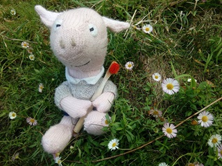 A small knitted piglet soft toy is sitting on grass, surrounded by dandelions.