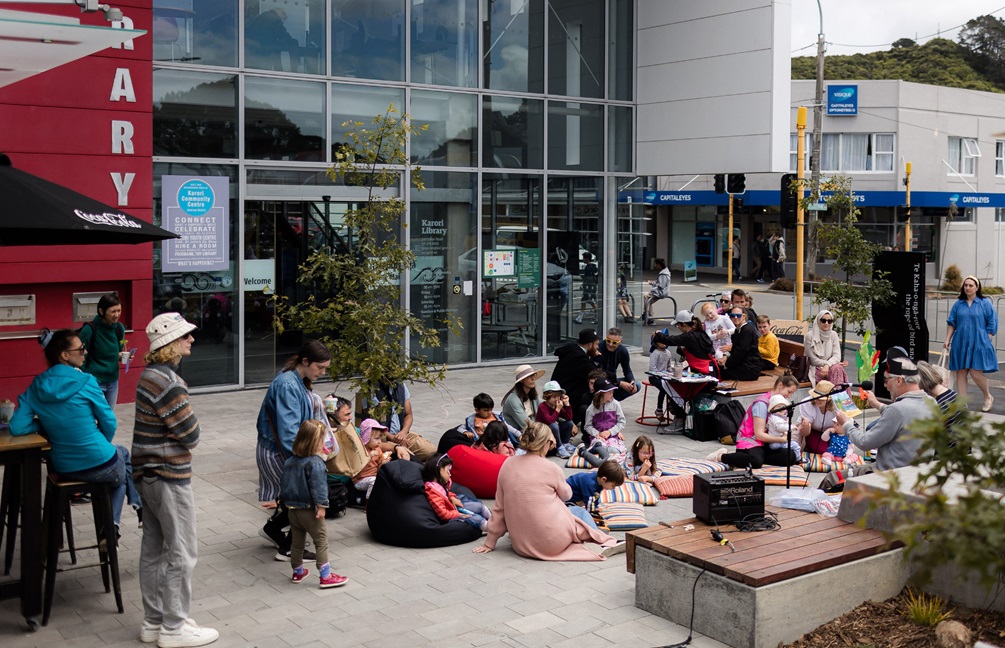 A group of people, standing and sitting in front of the Karori Library entrance, enjoying storytelling at the Karori Town Centre upgrade celebration.