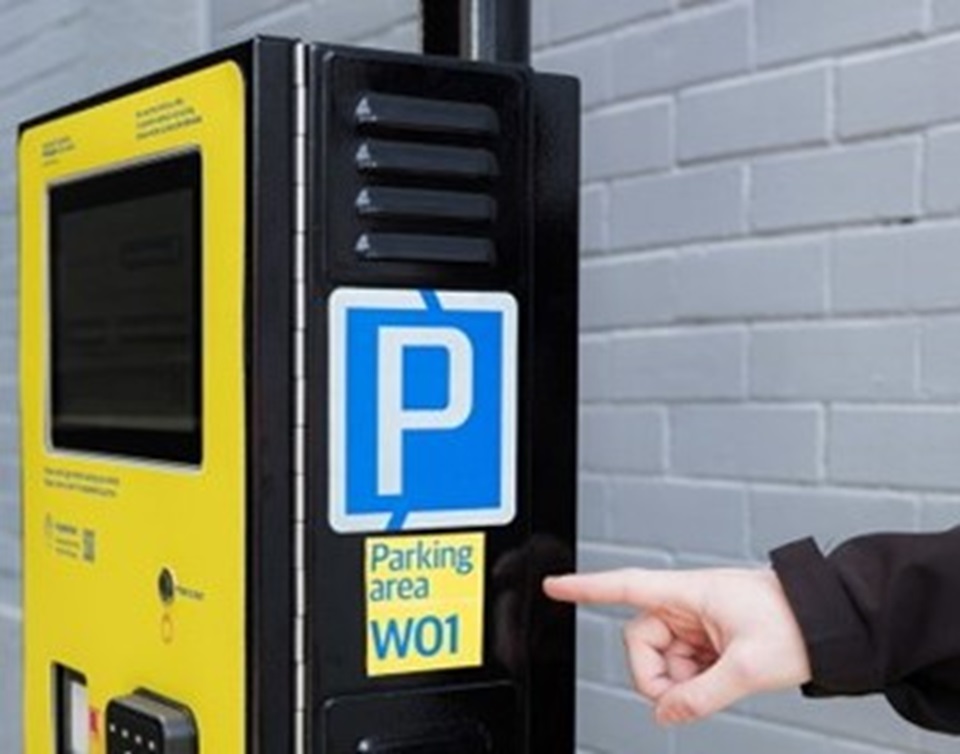 Pay by Plate parking meter with a person's hand pointing to parking area 'W01' on the side of the meter.
